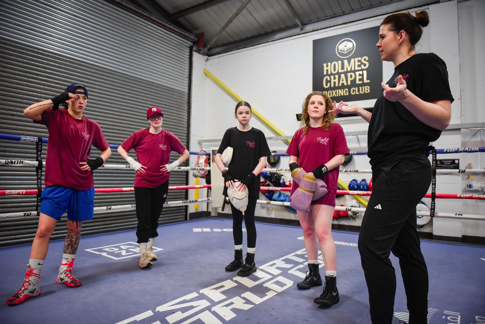 A coach talking to four girls standing in a boxing ring, two holding pads and gloves