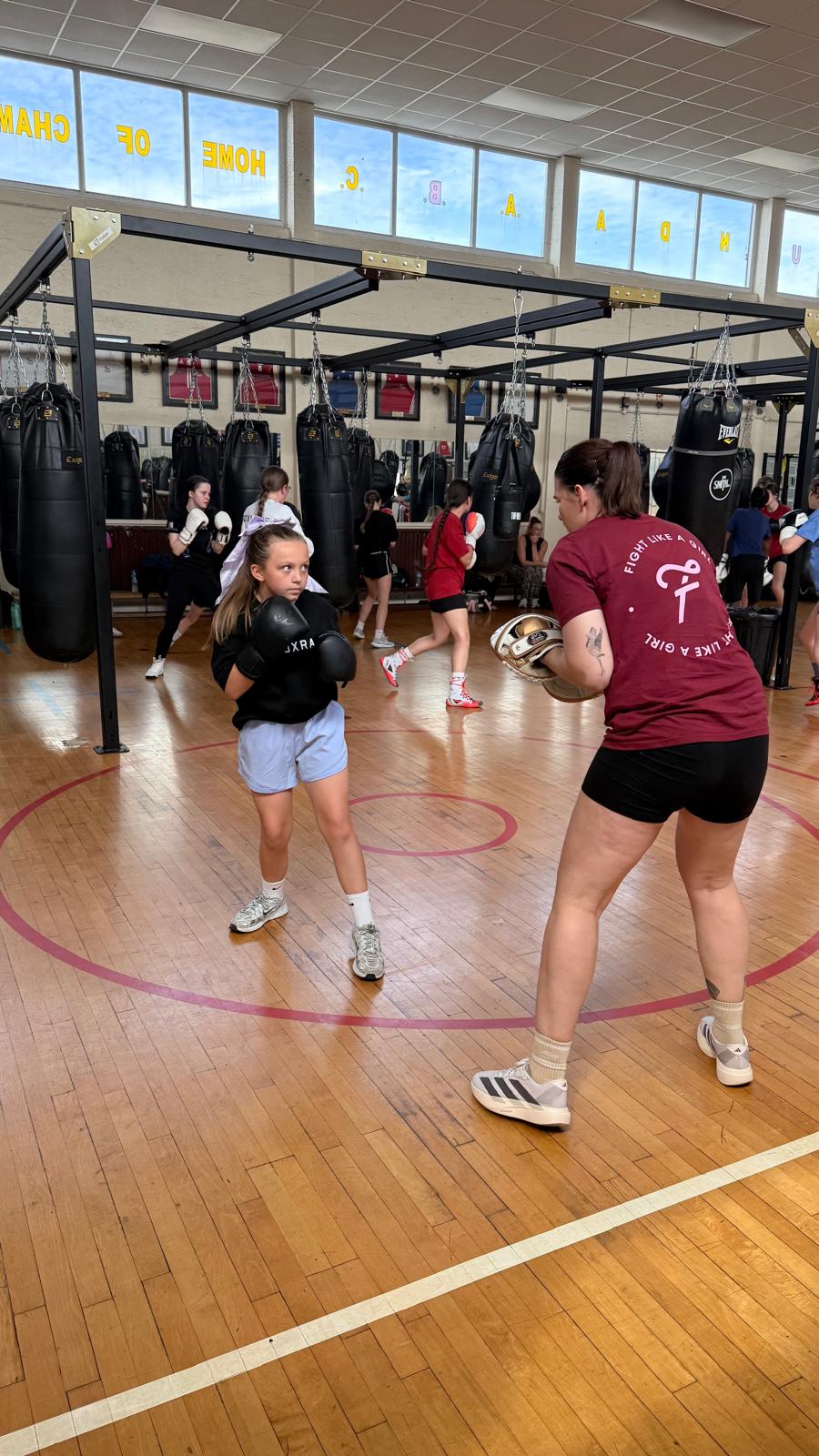 A girl on the pads with a coach in a busy boxing gym