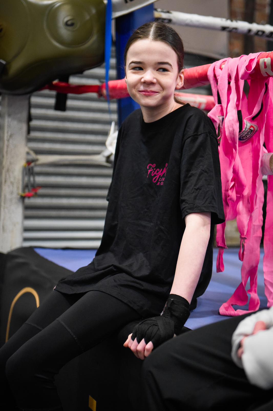 A smiling girl in a black Fight Like a Girl t-shirt with hand wraps, sitting on the edge of a boxing ring