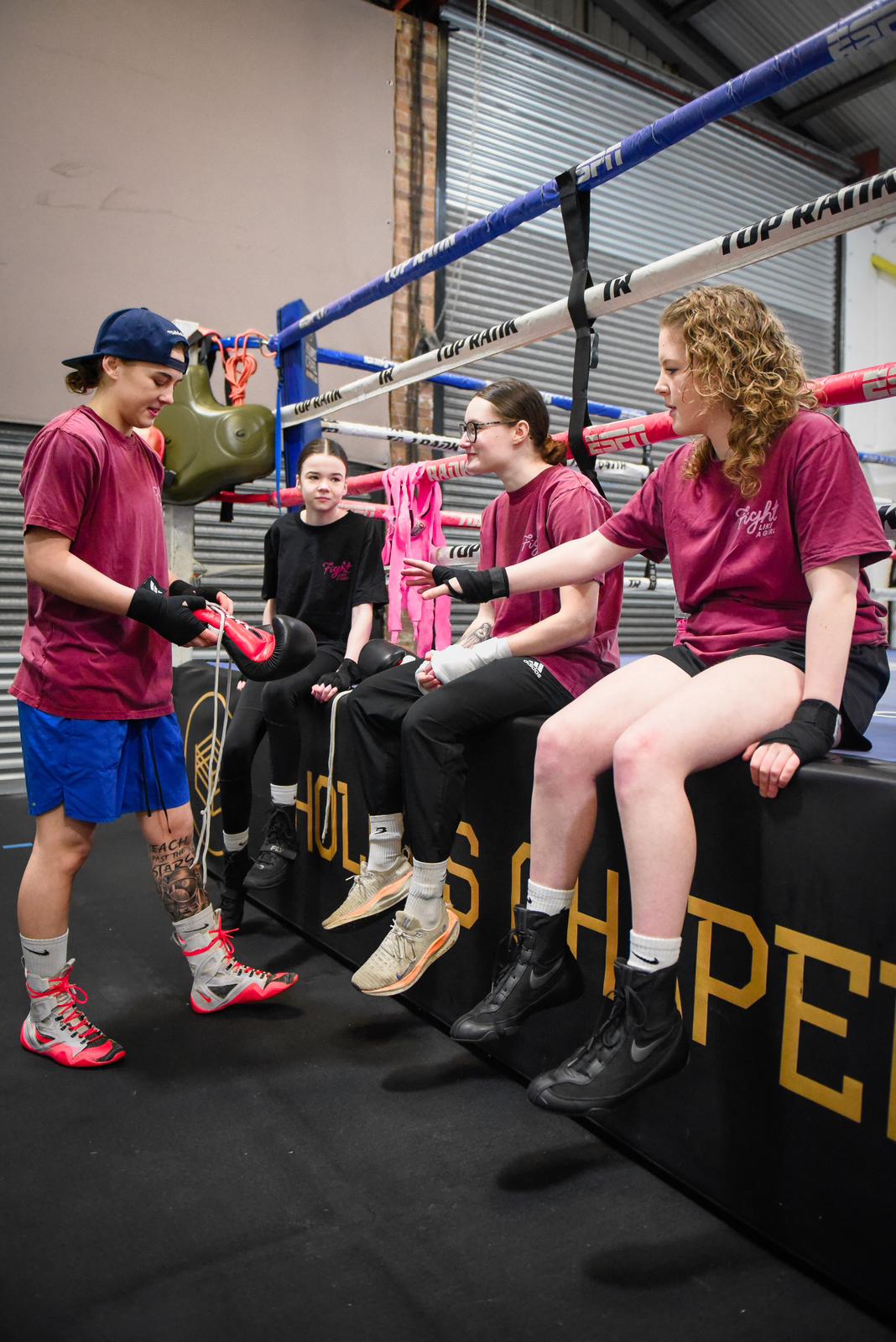 Four girls in Fight Like a Girl t-shirts sitting on the edge of a boxing ring while a coach laces a glove
